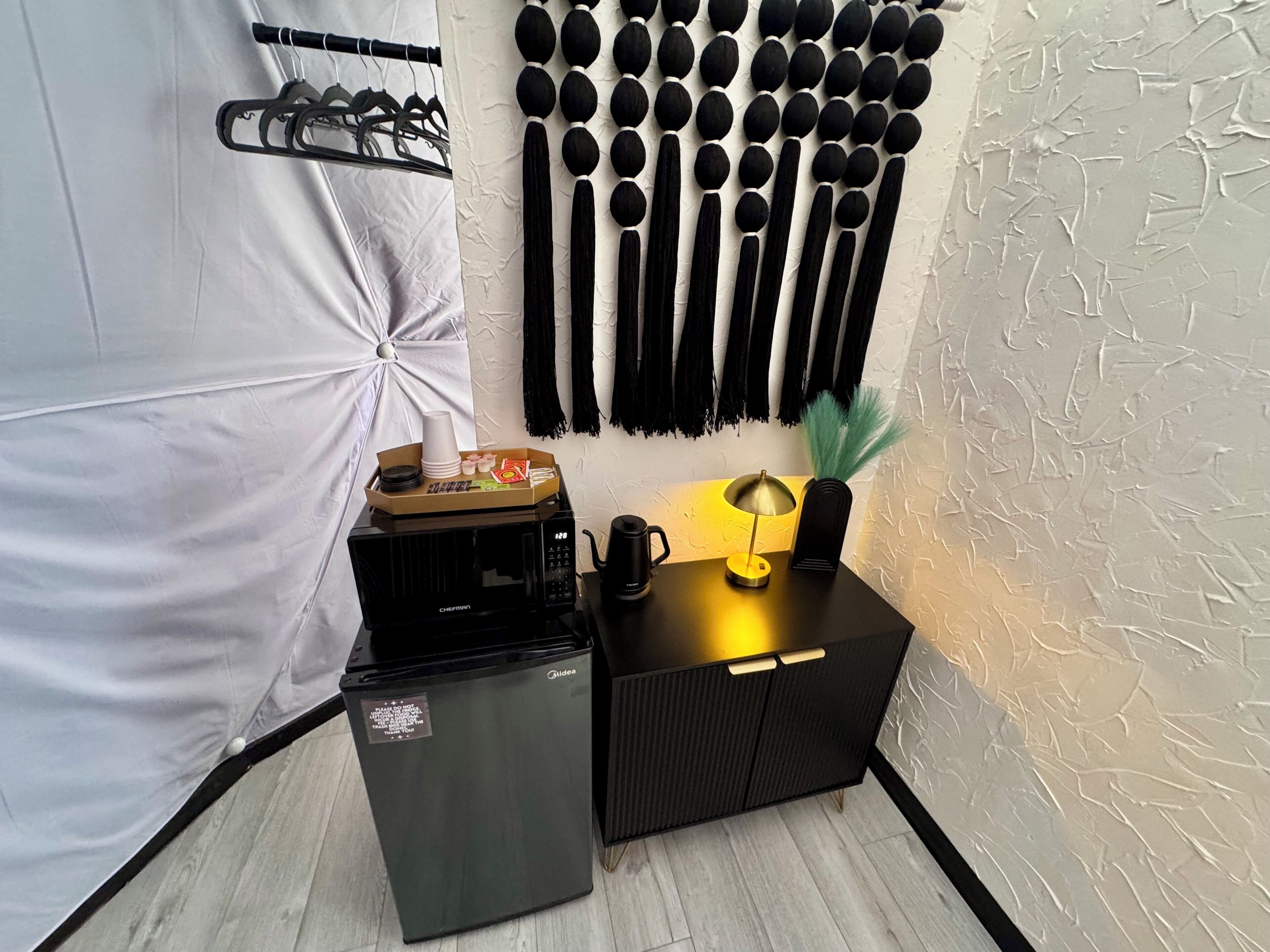 A kitchenette with a microwave, a small refrigerator, and a coffee maker on a black countertop. There is a tray with cups and snacks on the microwave.