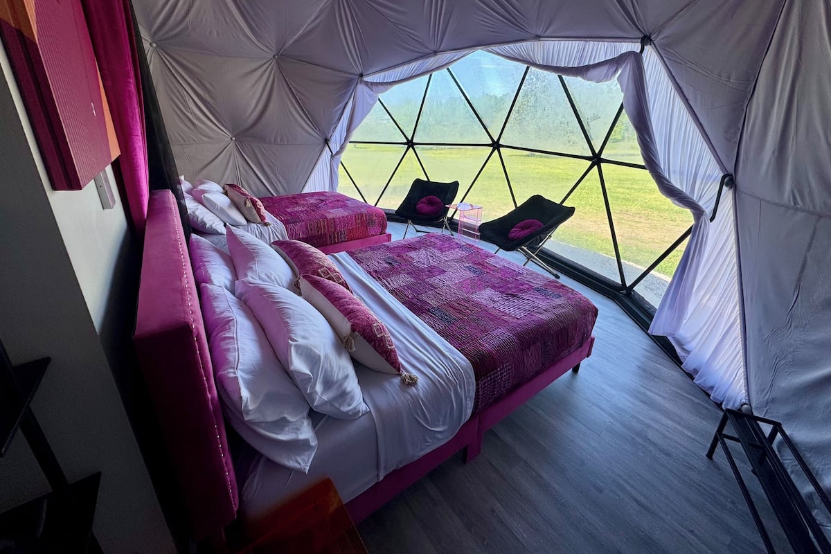 A tent-style bedroom with two beds, each with pink bedding and pillows, and a view of a grassy field through large windows.