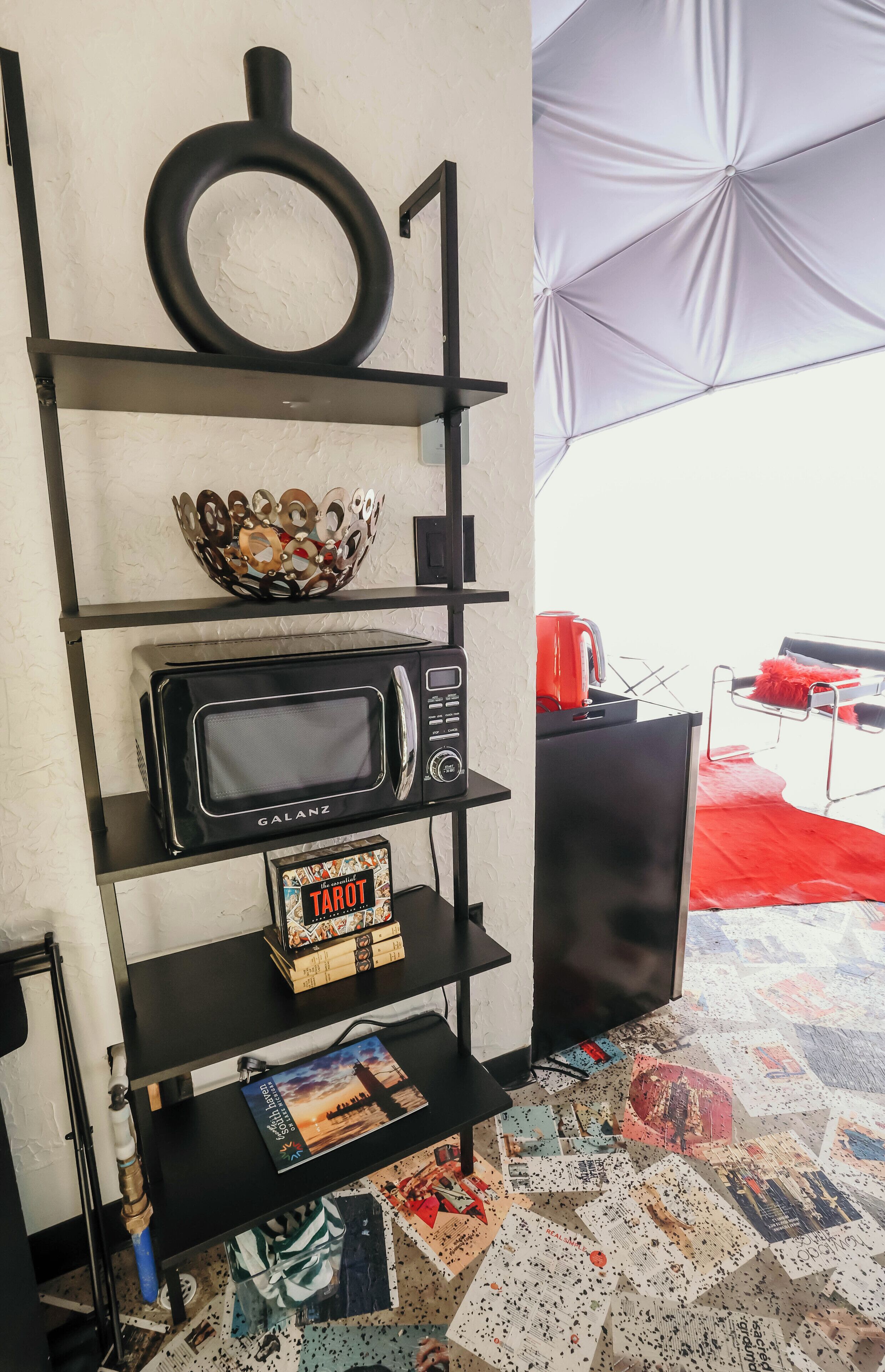 Una habitación con un estante negro que contiene un microondas, un cuenco decorativo y una caja de cartas del tarot.