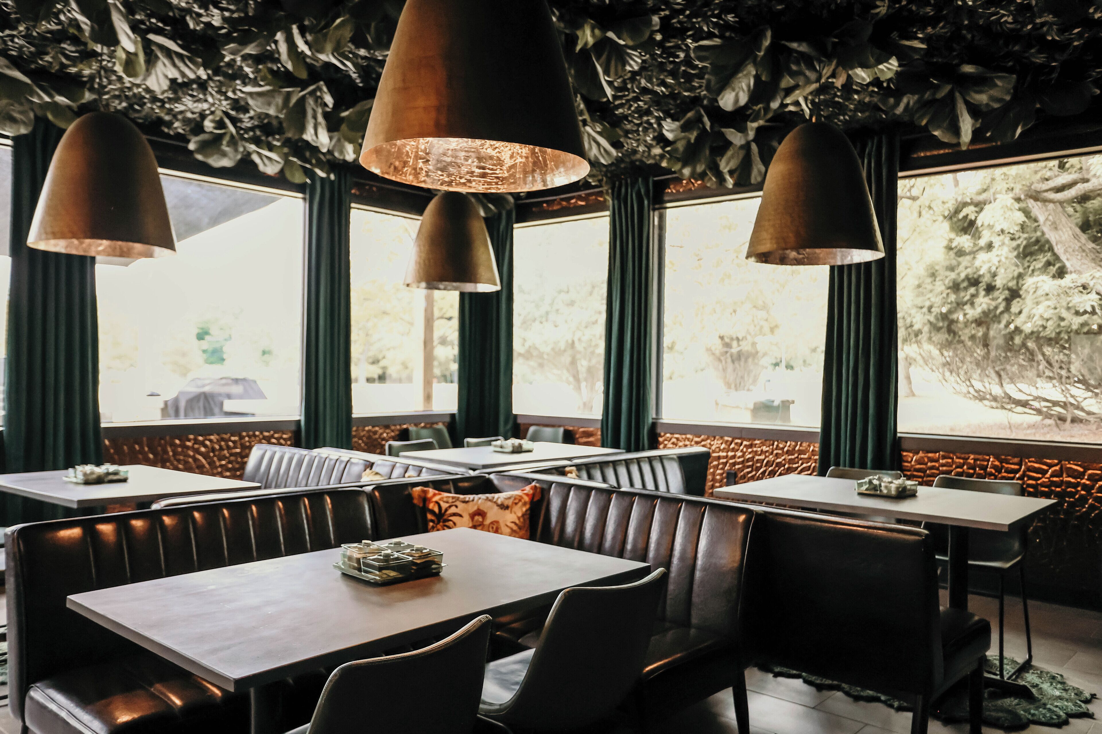 L’intérieur d’un restaurant avec des banquettes, des luminaires suspendus et de grandes fenêtres donnant sur un paysage arboré.