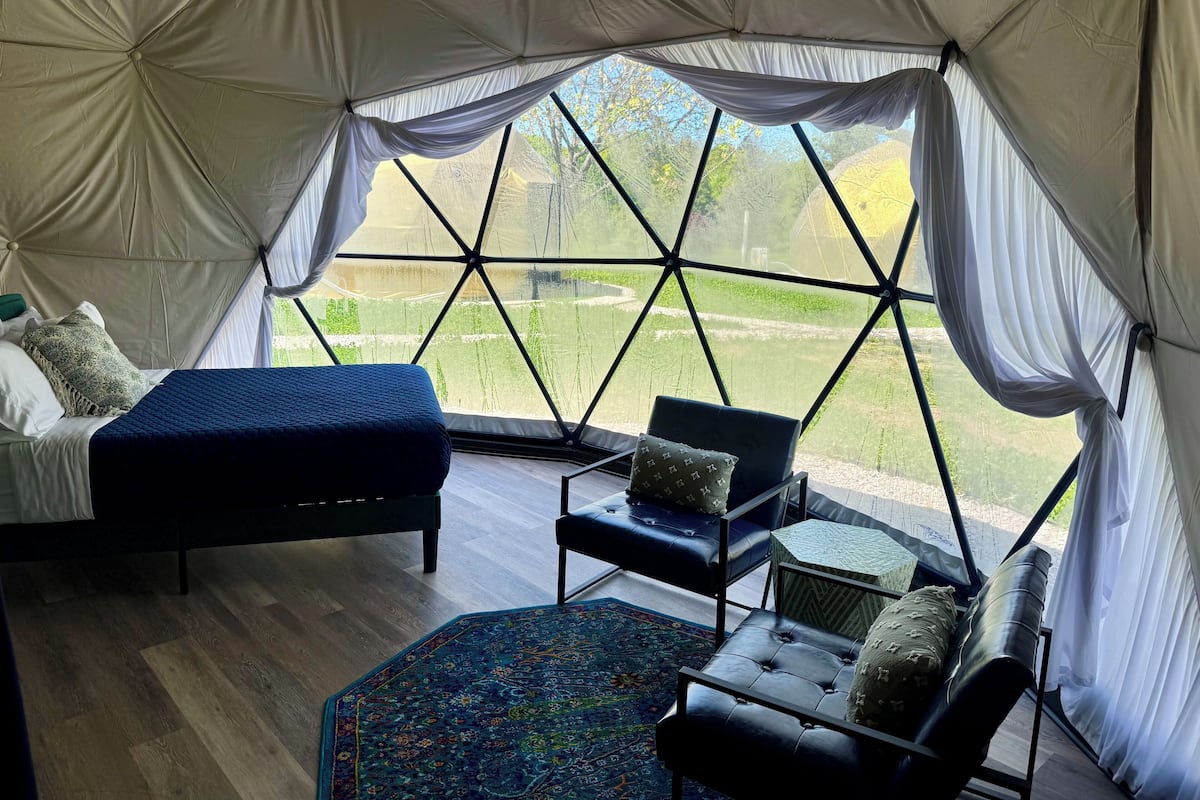 A tented room with a bed, two chairs, a small table, and a view of a green landscape.