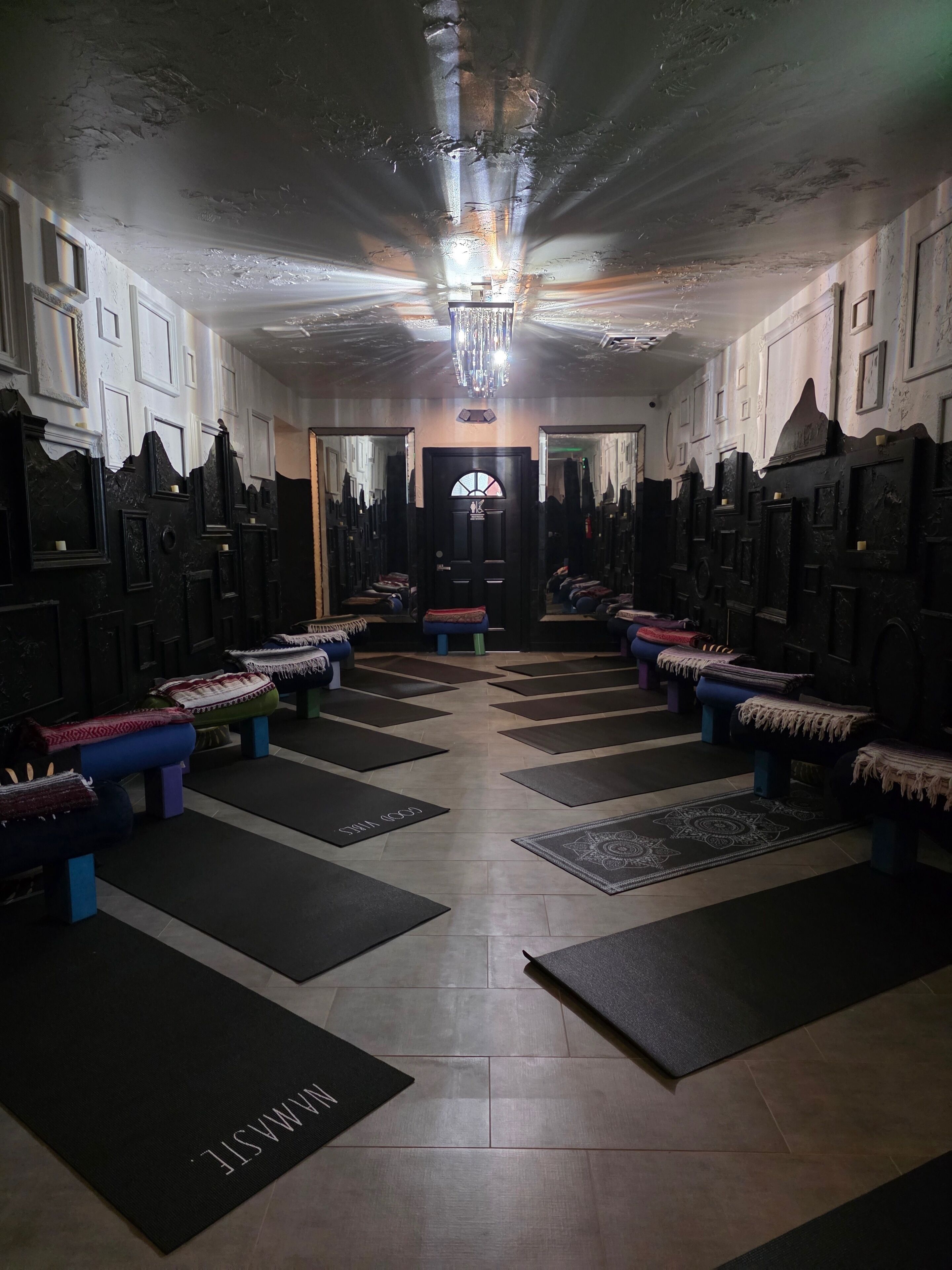 A hallway with benches and mats, leading to a door with a mirror and a chandelier.
