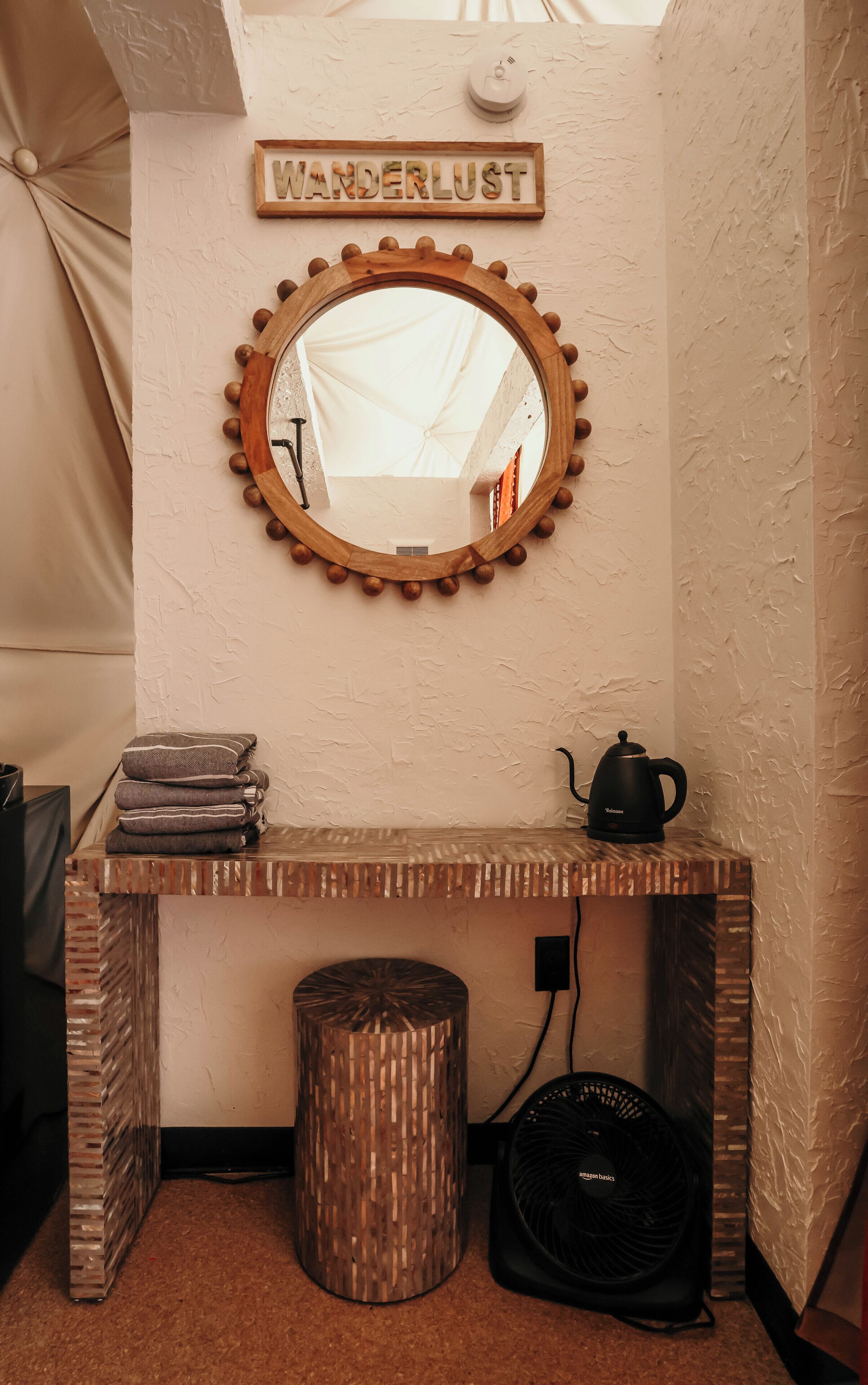 Une petite pièce comprenant une coiffeuse en bois, un miroir rond encadré en bois, un petit tabouret et une étagère murale où sont pliés des serviettes.