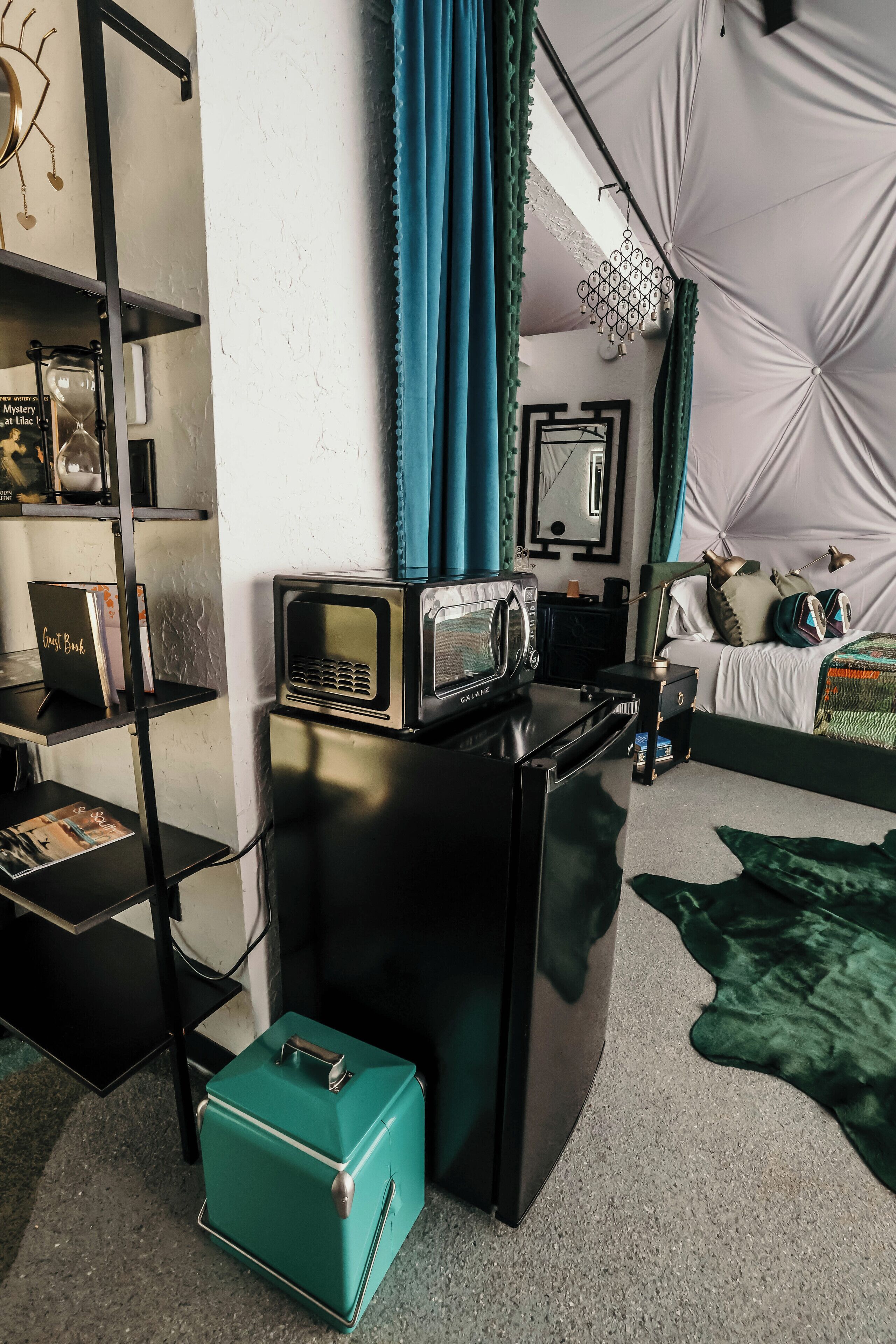 Una habitación pequeña con kitchenette, una cama, una alfombra verde y una estantería.
