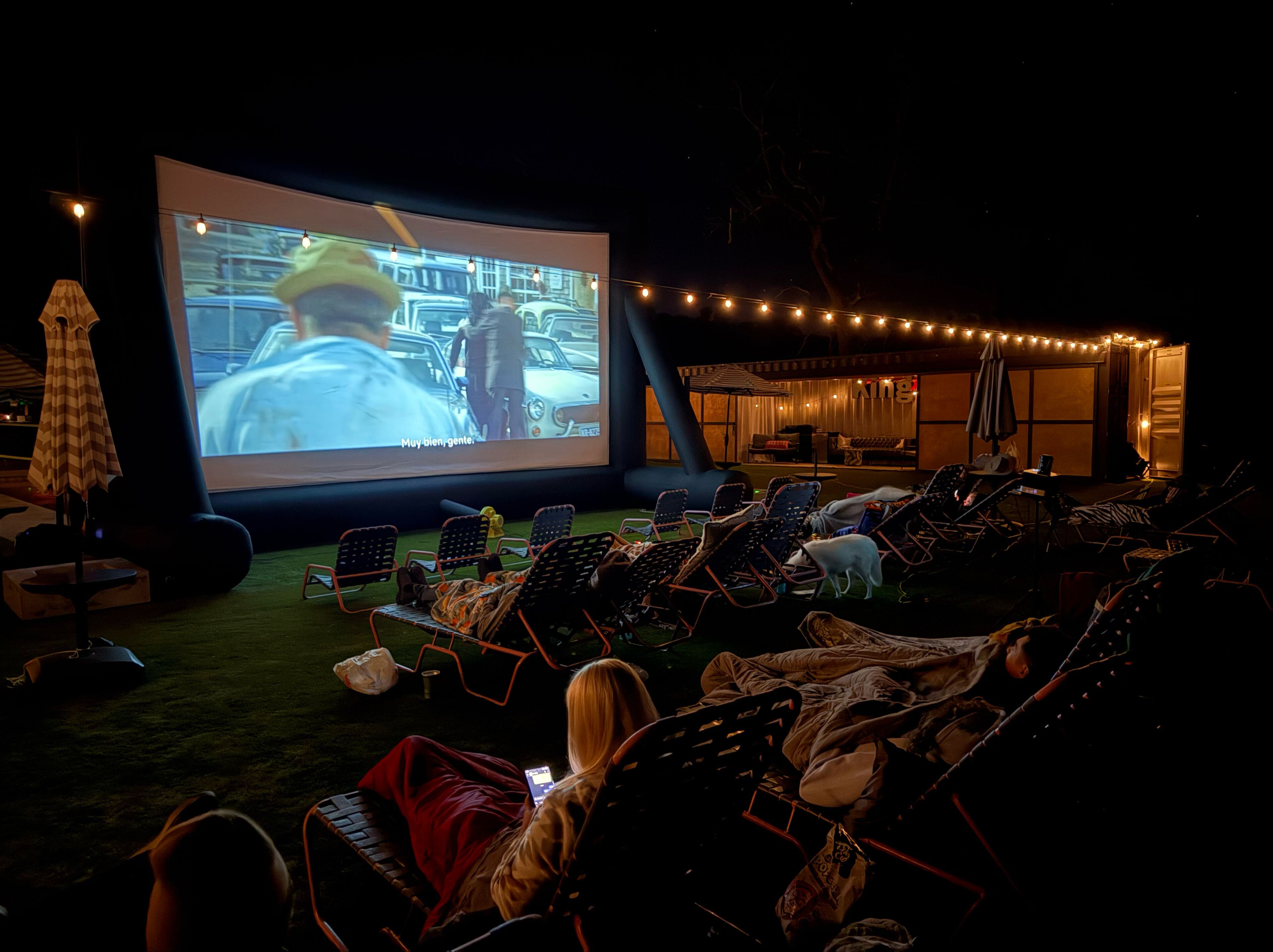 An outdoor movie screening area with a large screen, reclining chairs, and string lights.