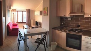 A compact kitchen with a stove, oven, and modern cabinetry. There is a dining area with a table and chairs, and a red sofa in the background. - Case Cosi Verona - Appartamento 1-4 (Verona)