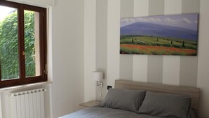 A bedroom with a bed, a nightstand, a lamp, a window with a view of greenery, and a painting on the wall. - Case Cosi Verona - Appartamento 1-4 (Verona)