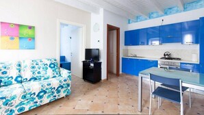 Un espacio habitable compacto con sofá, televisor, mesa de comedor y zona de cocina con armarios azules.