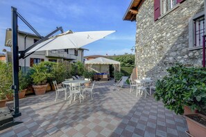 Un patio al aire libre con suelo a cuadros, mesas y sillas blancas, un paraguas blanco y macetas con plantas.