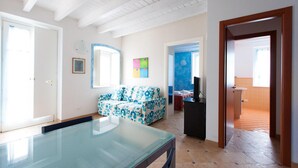 Un salón espacioso con una mesa de centro de vidrio, un sofá azul con estampado floral y un televisor. Se ve una puerta que da a un balcón con piso naranja.