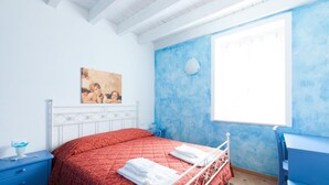 Un dormitorio con colcha roja, cabecera blanca, un cuadro en la pared, una ventana con cortinas y una silla azul.