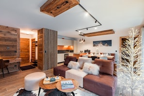 Un salon moderne avec un canapé, un pouf et une table basse, une cuisine en arrière-plan et un plafond en bois.