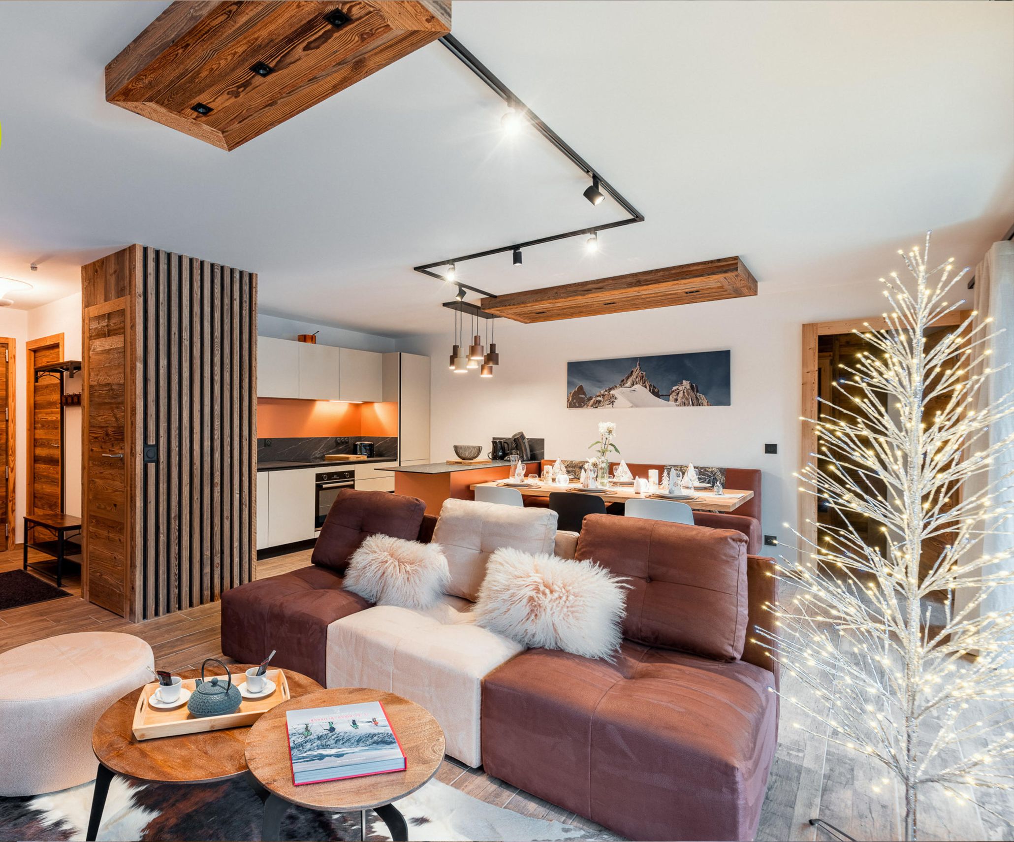 Un interno moderno con soffitto in legno, una zona salotto accogliente con divano e poggiapiedi, un tavolo da pranzo e una cucina sullo sfondo.