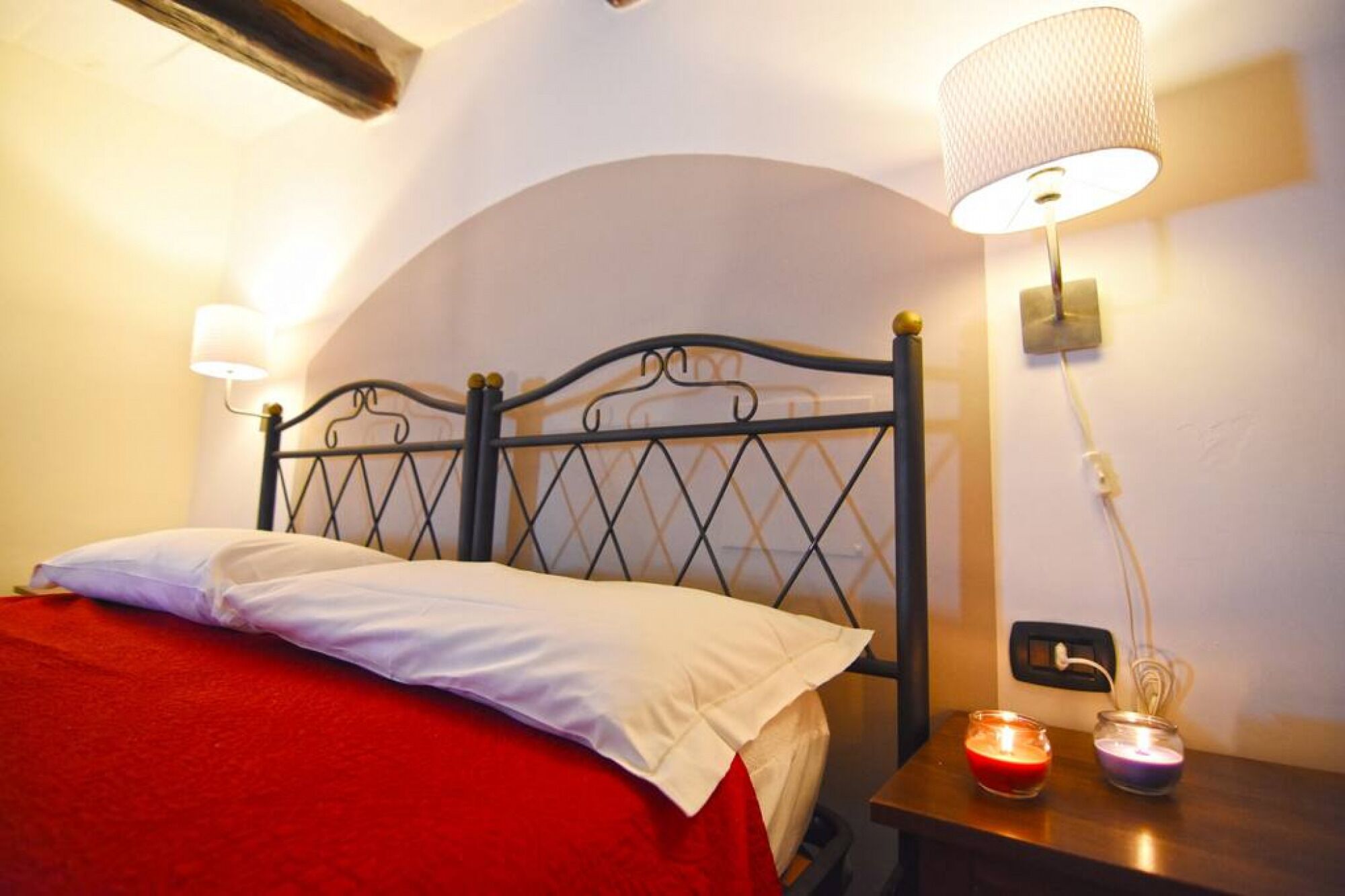 A bed with a red blanket and white pillows, a metal headboard, a wall-mounted lamp, and two lit candles on a bedside table.