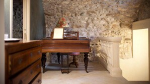 Een kamer met een stenen muur, een houten piano, een tafeltje met een lamp en een bank.