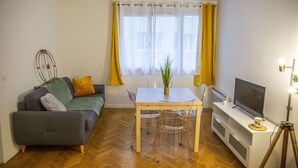 Un espace de vie compact comprenant un canapé, une table à manger avec des chaises, un téléviseur posé sur un meuble et une plante en pot.