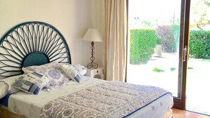 Un dormitorio con cama, mesita de noche, lámpara, ventana con cortinas y vistas a la vegetación.