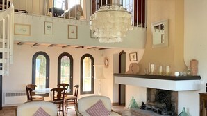 Amplia sala de estar con techo alto, chimenea y candelabro. Hay dos sillones, una mesa de centro y una mesa de comedor con sillas.
