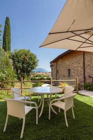 A patio with a round table and four wicker chairs, a large beige umbrella, and a stone building in the background. - Barchi Resort Apartments Suites Villa Castello - Loft Villa Castello (San Felice del Benaco)