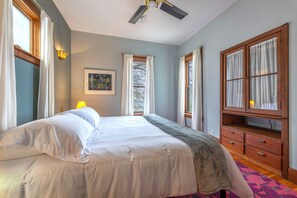 Un dormitorio con cama, mesita de noche, ventana con cortinas, ventilador de techo y un armario de madera.