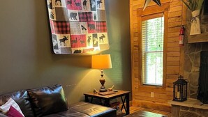 A cozy living room with a leather sofa, a quilted wall hanging, a table with a lamp, and a stone fireplace. - Hawks Ridge Cabin (Saint James)