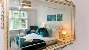 Un dormitorio con un espejo grande, un cabecero capitonado, una cama con ropa de cama color aguamarina, una lámpara de noche y una obra de arte en la pared.
