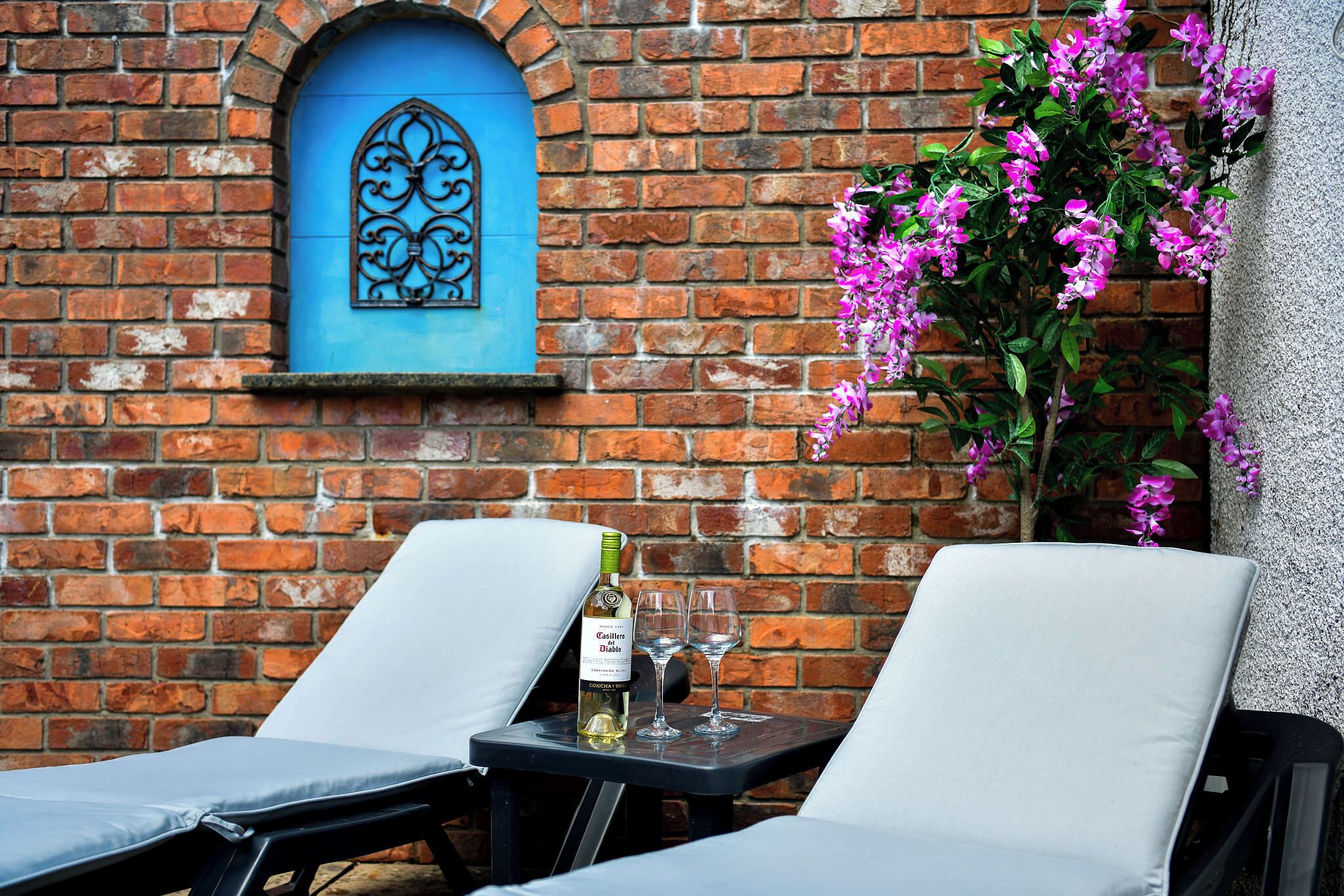 A brick wall with a blue door and a decorative metal piece. There are two lounge chairs and a table with a bottle of wine and two glasses.