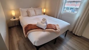 Un lit bien fait, avec des draps blancs, un plaid marron et une serviette pliée posée sur le lit. Deux lampes de chevet sont placées de chaque côté du lit.