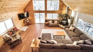 A cozy cabin interior with a high ceiling, wooden walls, a stone fireplace, a large window, and a television.
