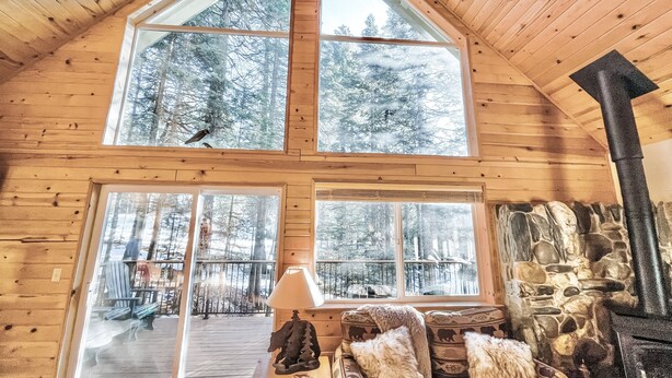 A cabin interior with a high ceiling, wooden walls, a stone fireplace, and large windows offering a view of a snowy forest.