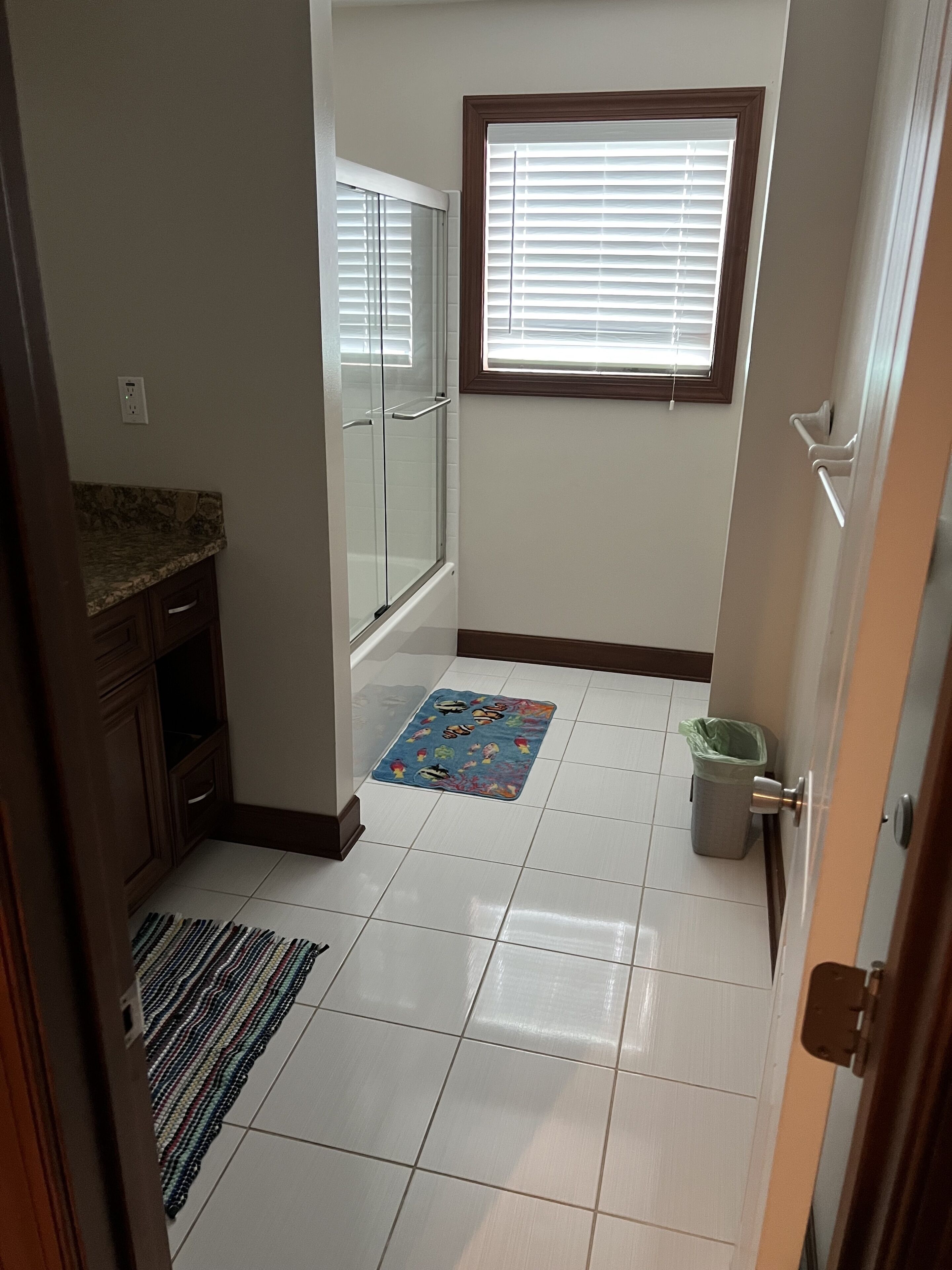 A bathroom with a shower, a window with blinds, a tiled floor, and a small rug.
