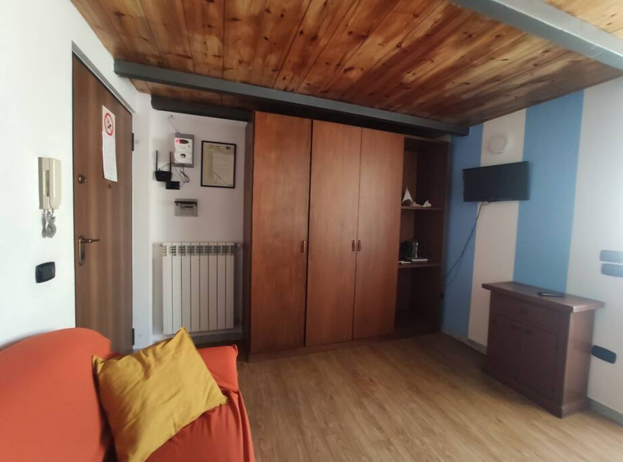A room with a wooden ceiling, a radiator, a wardrobe, a TV mounted on the wall, and a small desk.