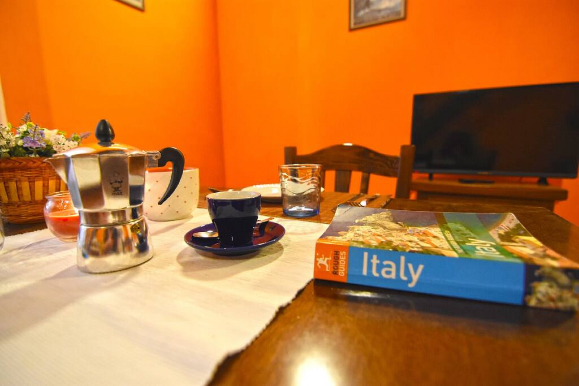 A dining table with a coffee pot, a cup, a glass, and a book titled "Italy" on a tablecloth.