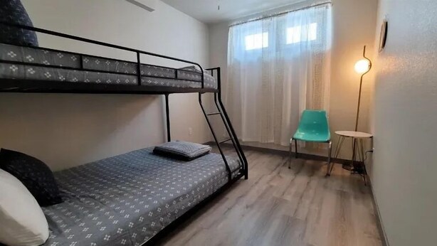 A bunk bed with a ladder, a small table, a chair, and a lamp in a room with wooden flooring and a window with curtains.