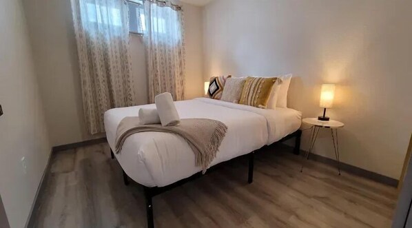 A neatly made bed with a blanket and pillows, a bedside lamp, and a window with curtains.