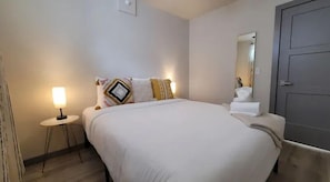 A neatly made bed with white bedding, a bedside lamp, and decorative pillows. A mirror and a towel stand are visible on the right side of the bed.