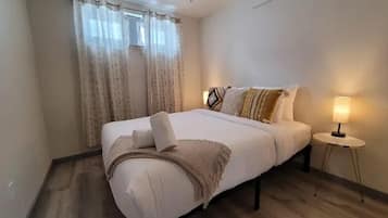A neatly made bed with a white comforter and pillows, a bedside lamp, and a window with curtains.