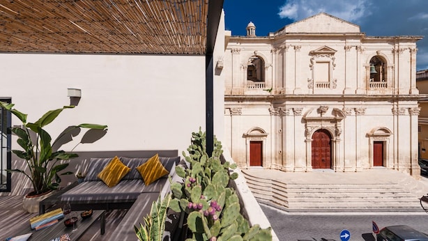 A modern outdoor seating area with a sofa and cushions, a potted plant, and a cactus. In the background, there is a historic building with a bell tower and a staircase leading to the entrance. - Casa San lio - Casa San Lio (Noto)