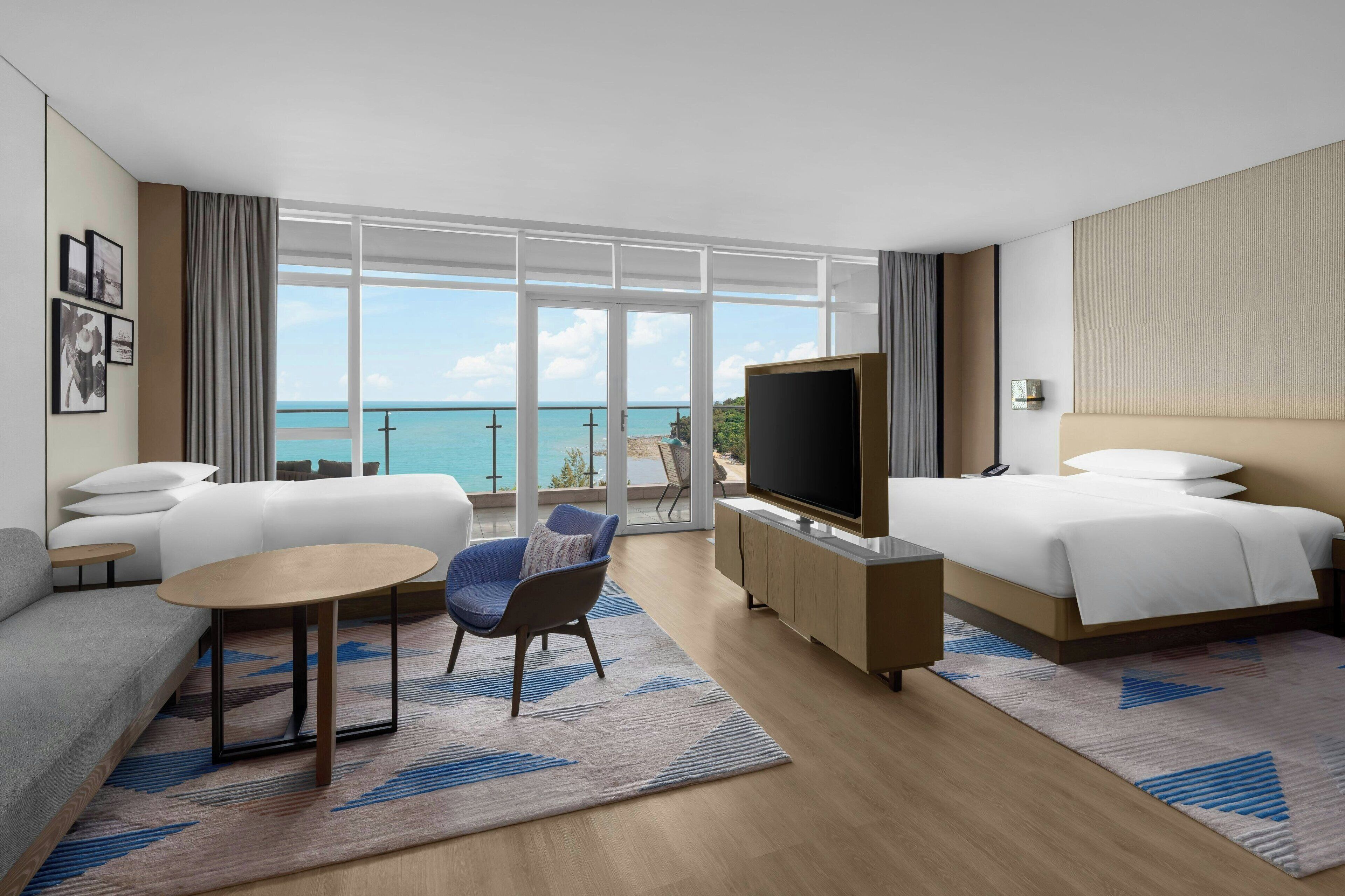 A modern hotel room with a large balcony, a sofa, a chair, a TV, and a bed.