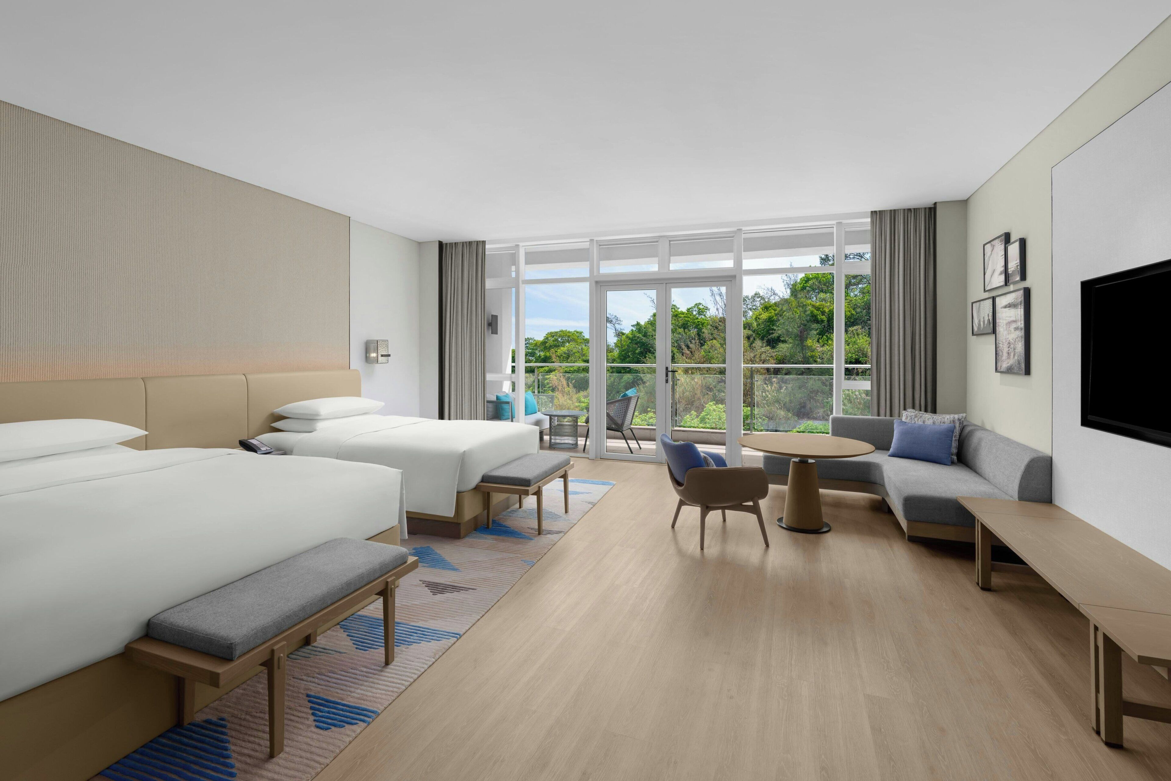 Moderni hotellihuone, jossa on suuri sänky, sohva, pieni pöytä ja parveke, jolta on näkymä vehreään maisemaan.