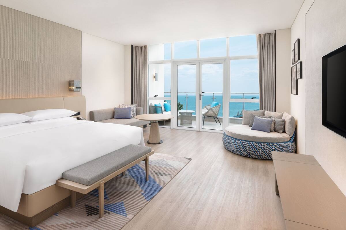 A modern hotel room with a large bed, a seating area with a view, and a balcony with a pool.