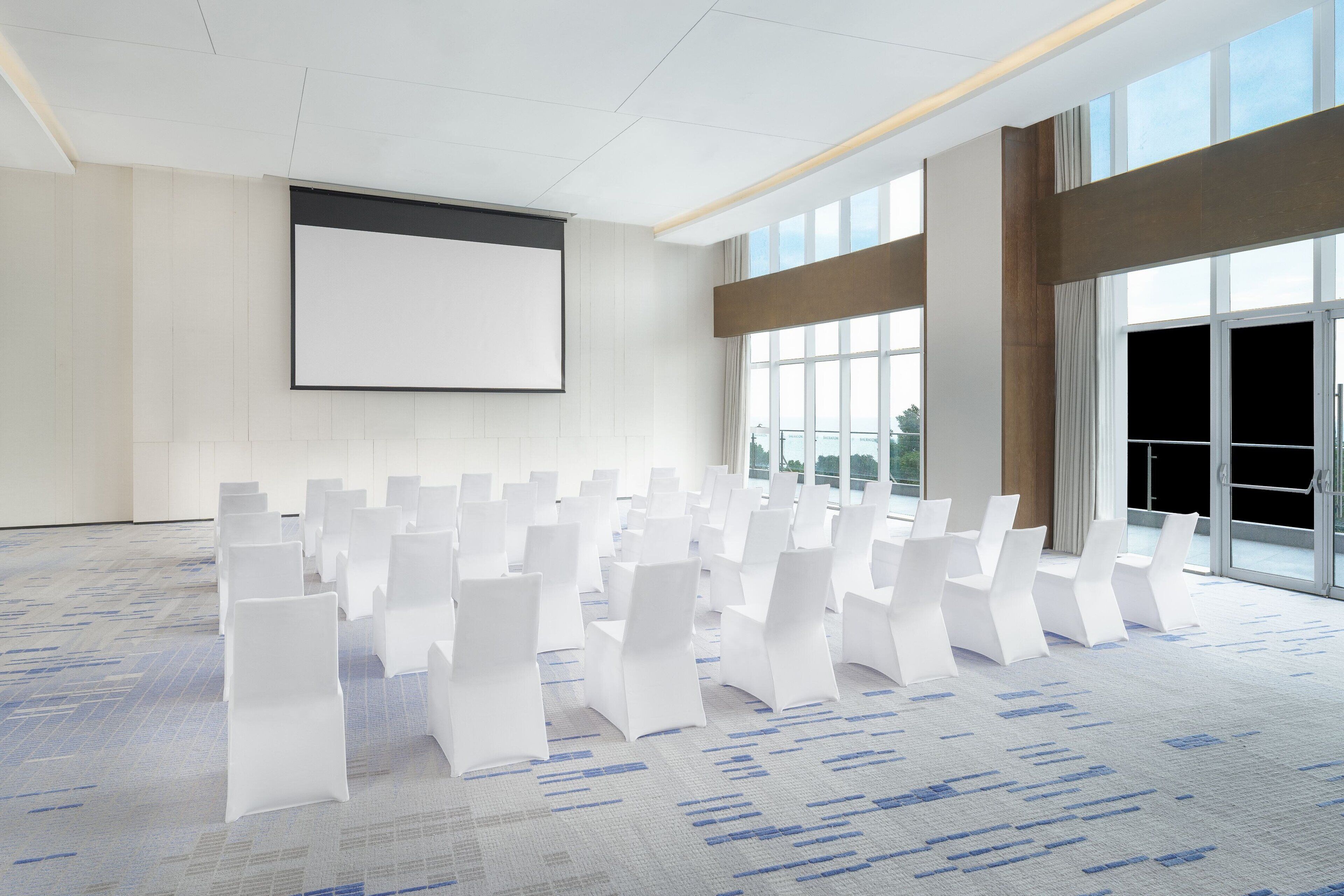 Uma sala de conferências com fileiras de cadeiras brancas, uma tela grande em branco e amplas janelas com vista para o exterior.