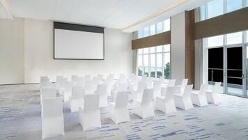 Uma sala de conferências com fileiras de cadeiras brancas, uma tela grande em branco e amplas janelas com vista para o exterior.