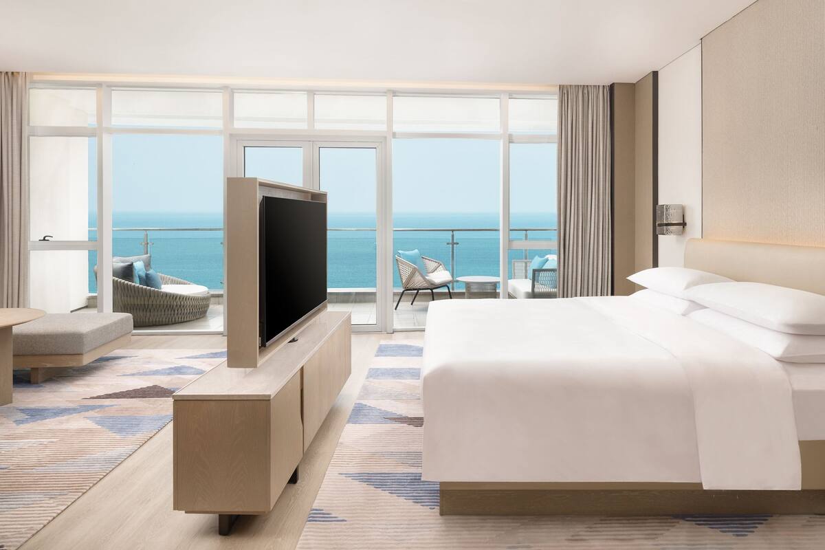 A modern hotel room with a large bed, a flat-screen TV, and a balcony with a view of the sea.