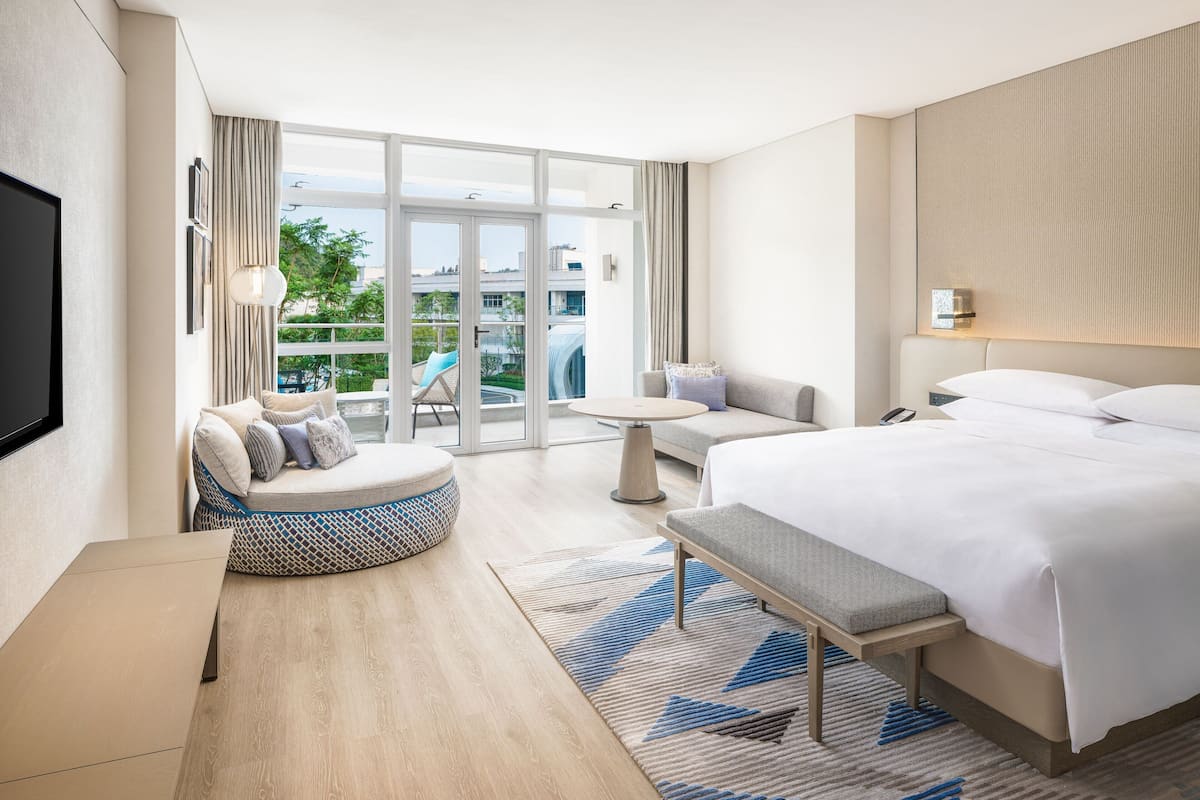A modern hotel room with a large bed, a seating area with a round table, and a balcony with a view of the city.
