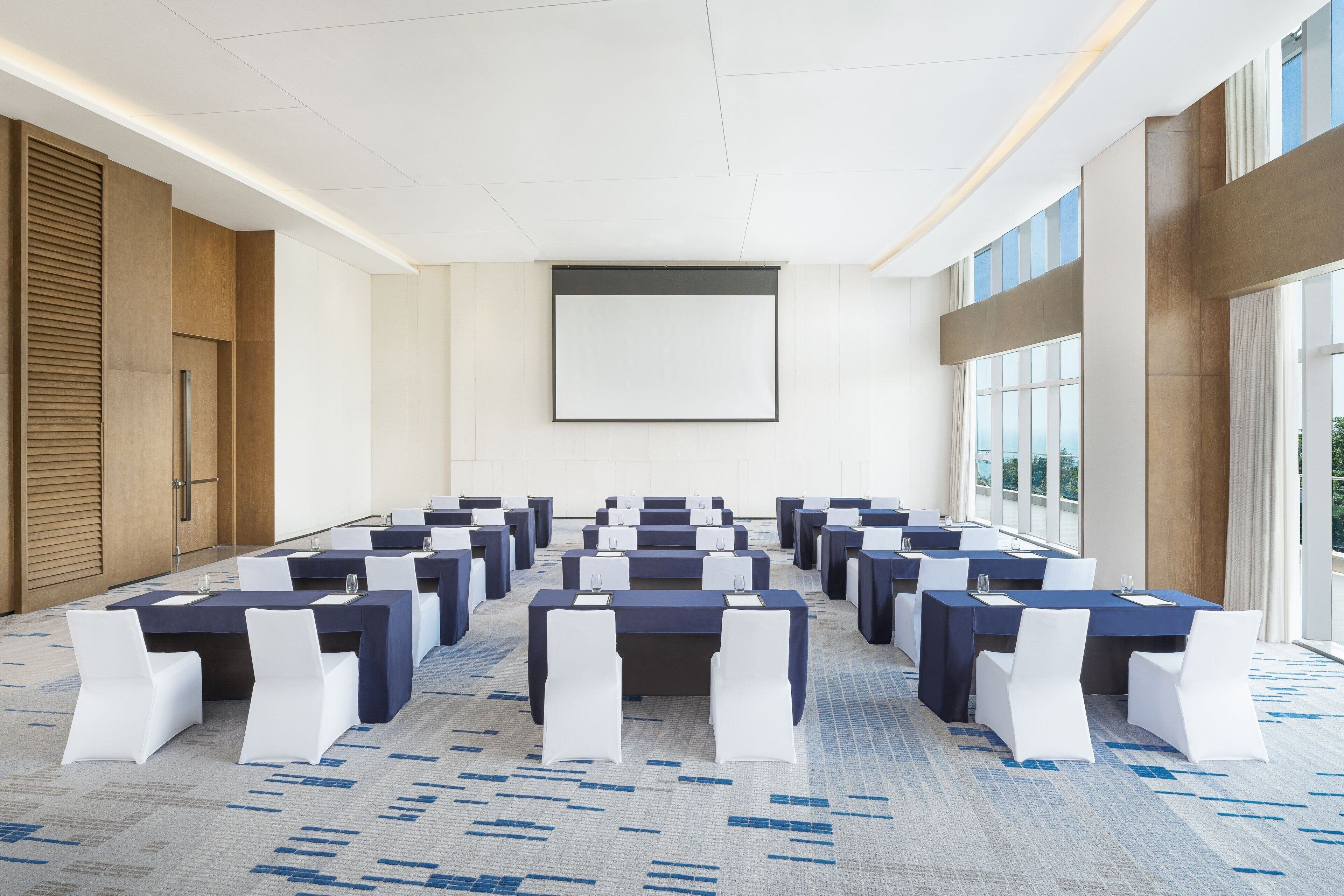 Een congreszaal met meerdere tafels en stoelen, grote ramen en een projectiescherm.
