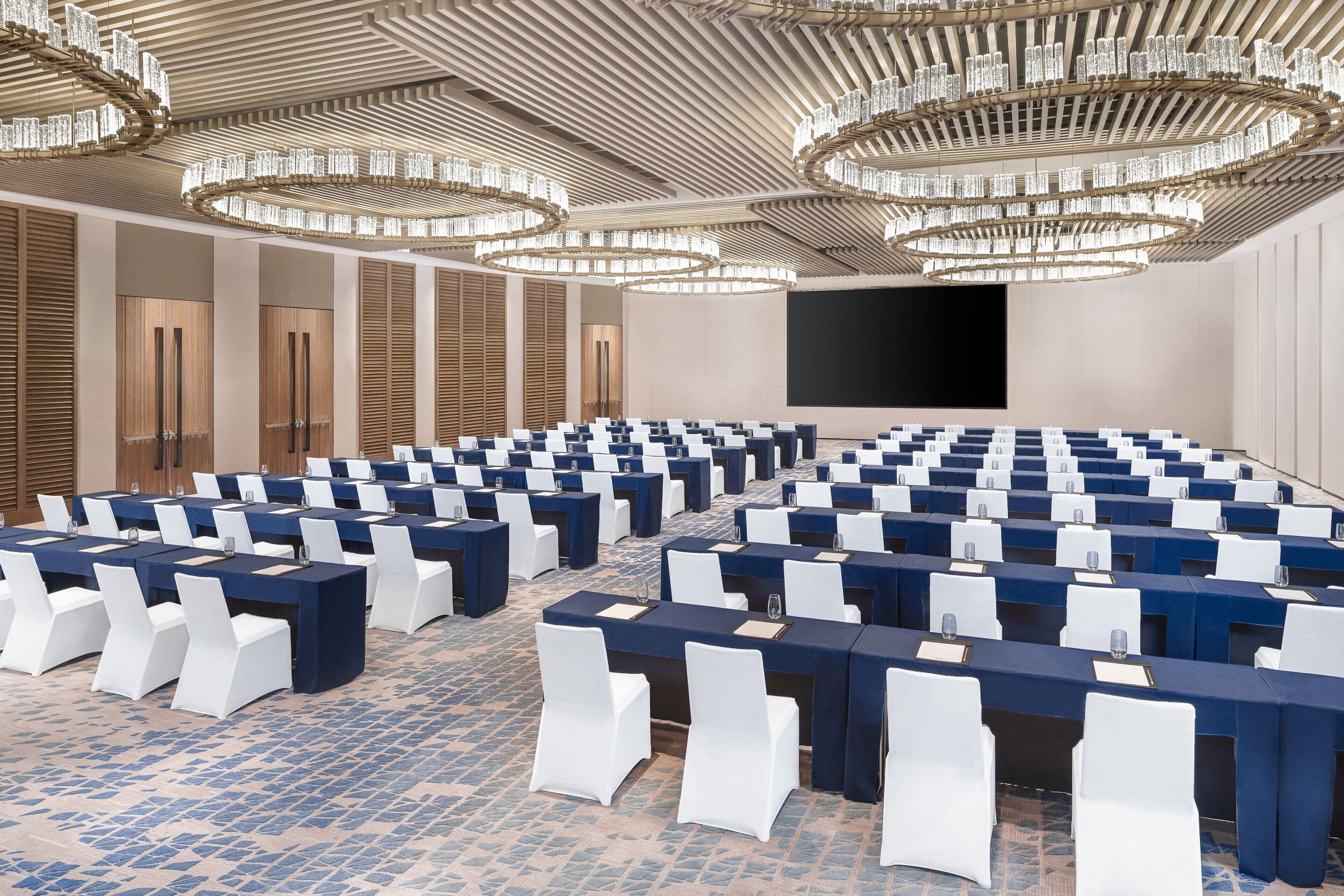 Un salón de conferencias con filas de mesas y sillas, una pantalla grande y una lámpara de araña moderna.