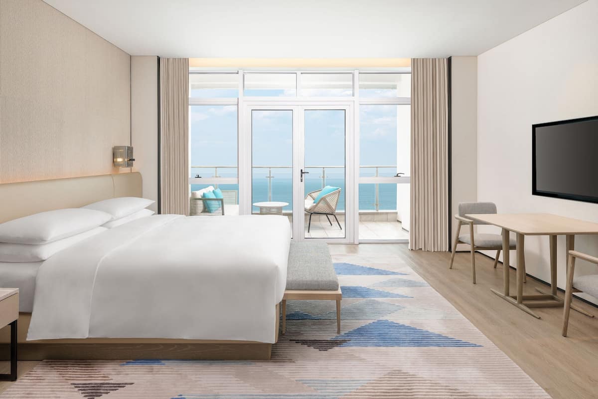 A modern hotel room with a large bed, a desk, and a balcony with a view of the sea.