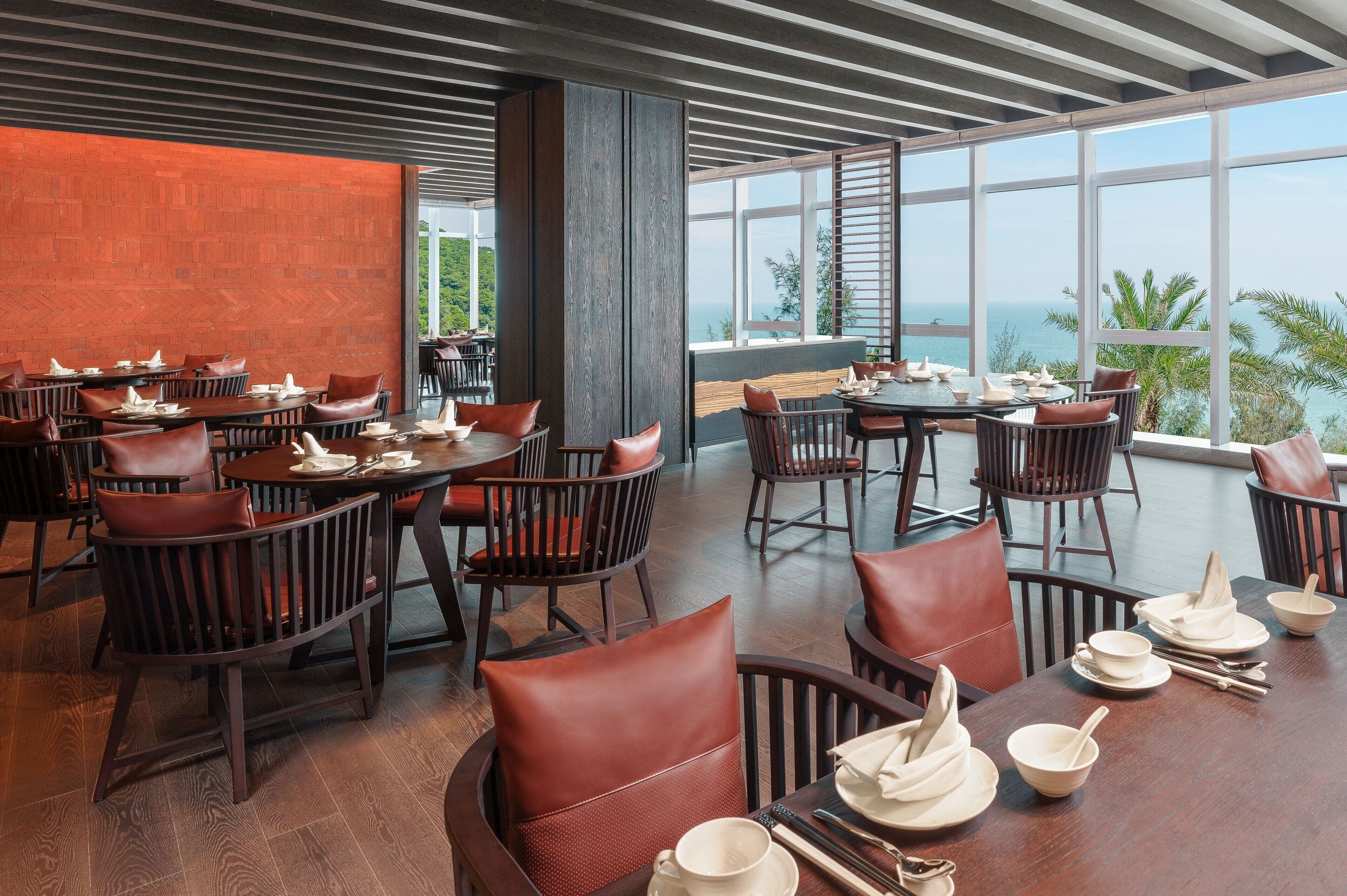 A restaurant with wooden tables and chairs, large windows with a view of the ocean, and a modern interior design.
