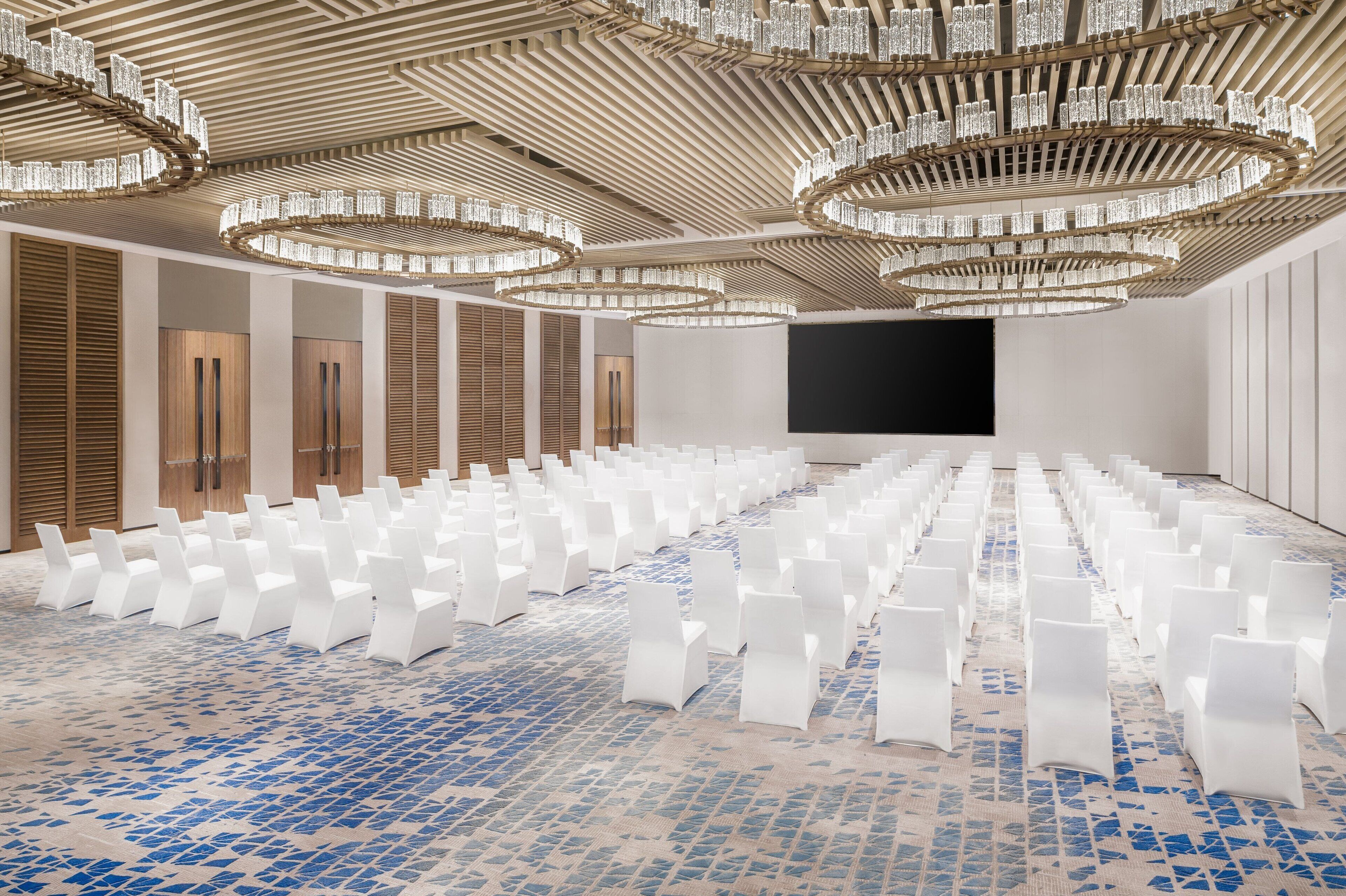 Un salón amplio con filas de sillas blancas, una pantalla grande y un suelo con diseño.